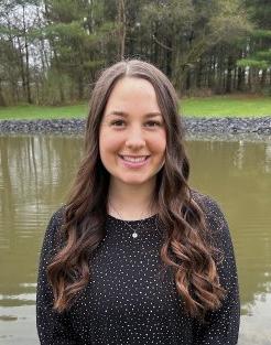 Sara Rachon, with long brown hair, stands in front of a body of water and smiles at the camera.