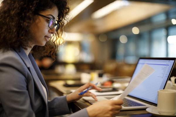 Woman in suit jacket and glasses holds a sheet of paper in one hand while typing on a laptop with the other hand.