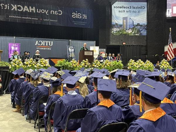 Members of the Class of 2024, in caps and gowns, sit in the Utica Memorial Auditorium as President Todd Pfannestiel speaks from a podium.